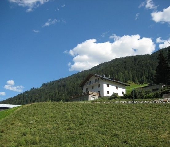 Ein weißes Haus befindet sich auf einem grasbewachsenen Hügel mit einer Bergkulisse, die von Bäumen bedeckt ist.