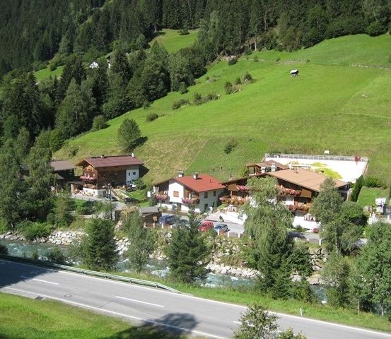 Luftaufnahme eines kleinen Dorfes in einem üppig grünen Tal, umgeben von Bäumen und einem mäandernden Fluss. Mehrere Häuser mit roten Dächern sind sichtbar, zusammen mit einer Straße und geparkten Autos.