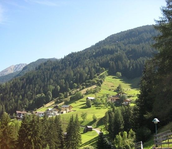 Ein Panoramablick auf eine Bergregion mit einer Ansammlung von Häusern, die zwischen üppigen grünen Feldern und dichten Wäldern unter einem klaren blauen Himmel eingebettet sind.