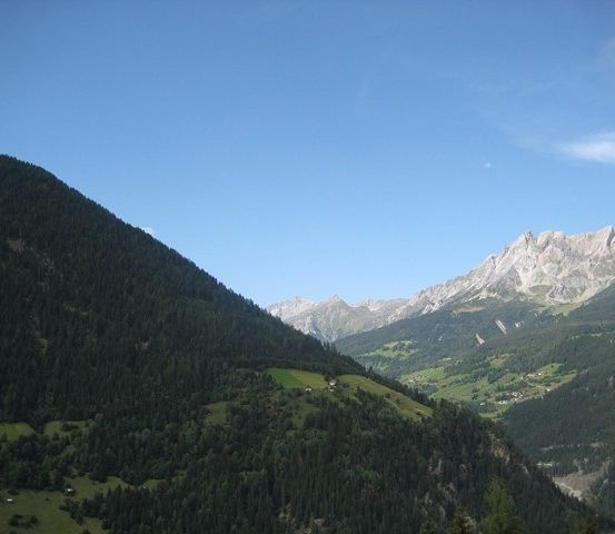 Ein Panoramablick auf eine Bergregion mit dichtem Wald und klarem blauem Himmel. Die Berge sind mit Bäumen bedeckt und haben einige grüne Grasflecken. In der Ferne gibt es einige Gebäude und einen Fluss.