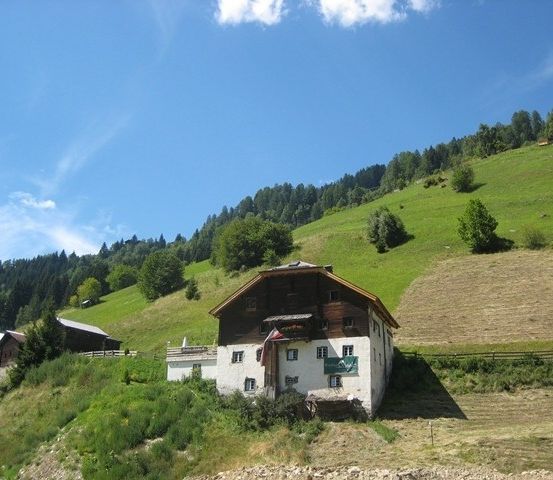 Ein rustikales Haus mit einer Fahne an der Seite befindet sich an einem Hang mit üppigem Grün und klarem blauem Himmel.