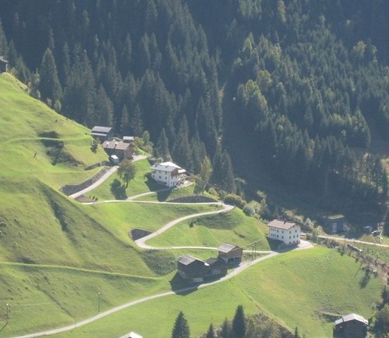 Ein Luftbild einer bergigen Region mit einer kurvenreichen Straße und mehreren kleinen Häusern, die zwischen Bäumen und grünen Feldern eingebettet sind.