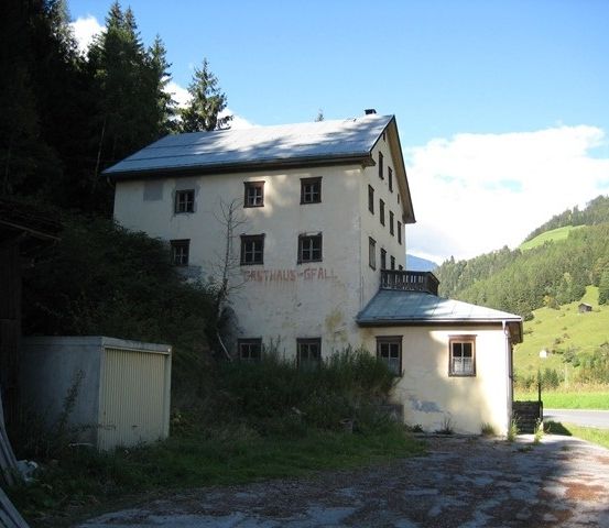 Ein altes, weißes Gebäude mit mehreren Fenstern und einem geneigten Dach, umgeben von Grün und Bergen.