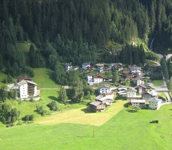 Eine Luftaufnahme eines kleinen Dorfes in einem Tal, umgeben von üppigen grünen Feldern und dichten Kiefernwäldern.
