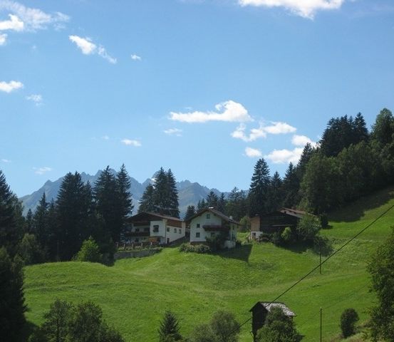 Ein Hügel mit einigen Häusern und einem kleinen Schuppen, umgeben von saftigem grünem Gras und Bäumen, unter einem klaren blauen Himmel mit verstreuten Wolken.
