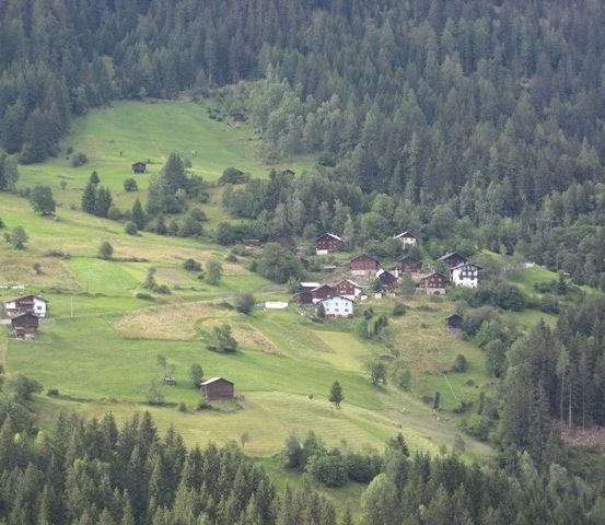 Eine Luftaufnahme eines kleinen Dorfes in den Bergen, umgeben von üppigen grünen Feldern und dichten Wäldern.