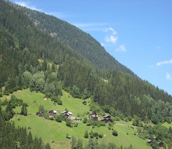 Ein Hügel mit kleinen Holzhäusern, umgeben von üppigem grünem Laub und Gras unter einem klaren blauen Himmel.