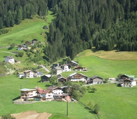Eine Luftaufnahme eines kleinen Dorfes, eingebettet in die üppigen grünen Berge, mit mehreren Häusern und einem Wald im Hintergrund.