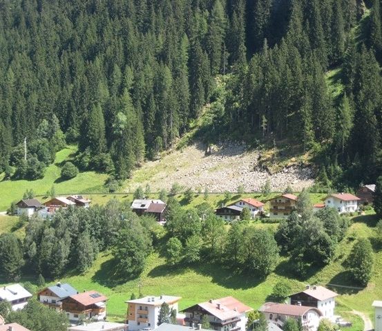 Luftaufnahme eines kleinen Dorfes in einem grünen Tal, umgeben von hohen Bäumen und Bergen. Die Häuser sind in verschiedenen Farben gestrichen.