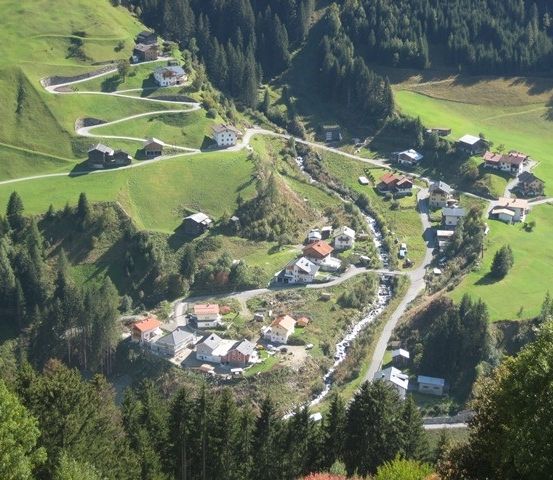 Eine Luftaufnahme eines kleinen Dorfes in einem grünen Tal, umgeben von Bäumen und einer kurvenreichen Straße.
