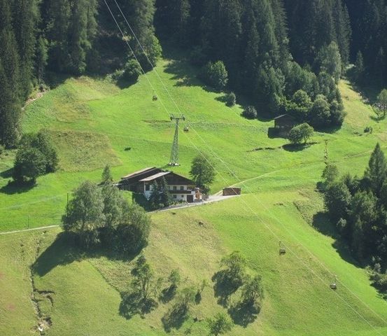 Eine Luftaufnahme eines grünen Tals mit einem Haus und einer Seilbahn. Das Haus liegt eingebettet in üppige Grünflächen, umgeben von Bäumen und Pflanzen. Die Seilbahn ist in der Ferne zu sehen, wobei Kabel das Tal überspannen.