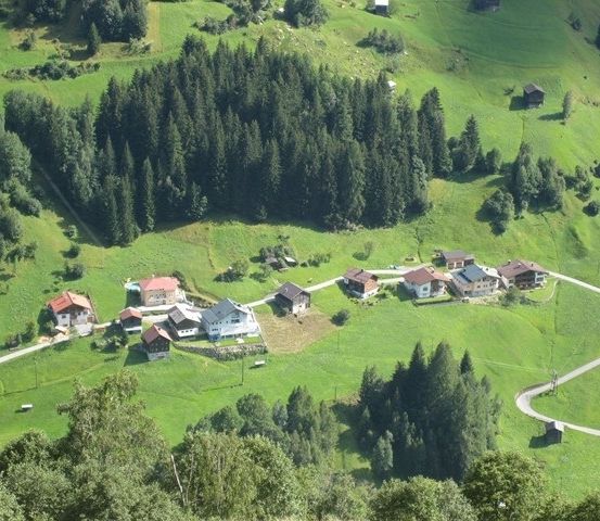 Eine Luftaufnahme eines kleinen Dorfes, eingebettet in einem üppigen grünen Tal, umgeben von dichten Wäldern und kurvenreichen Straßen.