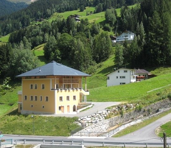 Ein gelbes Haus mit grauem Dach steht auf einem Hügel mit einer Straße davor. Es gibt zwei weitere Häuser am Hang, eines weiß und eines braun.