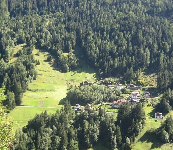 Luftaufnahme eines kleinen Dorfes, umgeben von üppigen grünen Feldern und dichten Wäldern. Das Dorf liegt in einem Tal mit einem klaren blauen Himmel darüber.
