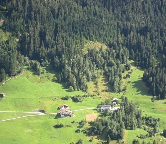 Eine Luftaufnahme eines friedlichen Tals mit üppigen grünen Feldern, dichten Wäldern und einigen verstreuten Häusern. Die Landschaft ist mit Bäumen und kleinen Gebäuden gespickt und schafft eine malerische Szene.