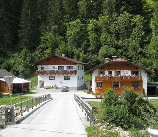 Zwei traditionelle Holzhäuser mit Balkonen, geschmückt mit Blumen, eingebettet in einer üppigen grünen Waldumgebung. Ein Auto ist vor einem der Häuser geparkt.