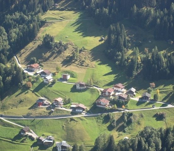 Eine Luftaufnahme eines kleinen Dorfes, eingebettet in einem Tal, umgeben von üppigen grünen Feldern und dichten Wäldern. Das Dorf besteht aus mehreren traditionellen Häusern mit roten Dächern, und eine kurvenreiche Straße verbindet sie.