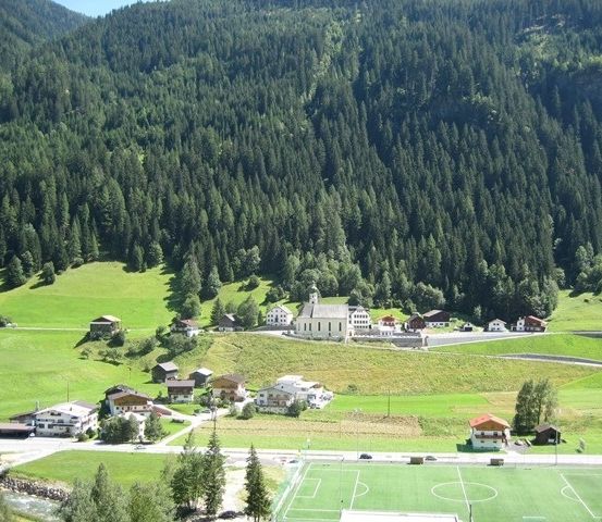 Luftaufnahme eines kleinen Dorfes in den Alpen mit einer Kirche, Häusern und einem Fußballfeld.