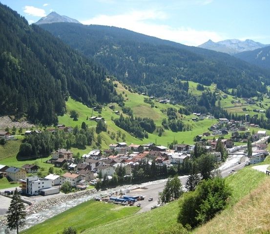 Luftaufnahme eines kleinen Dorfes in den österreichischen Alpen, umgeben von üppigen grünen Wiesen und dichten Wäldern. Das Dorf liegt an einem Hang mit einem Fluss in der Nähe. In der Ferne erheben sich majestätische Berge über der Landschaft.