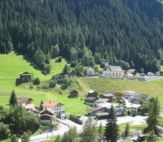 Luftaufnahme eines kleinen Dorfes in den österreichischen Alpen, umgeben von üppigen grünen Wiesen und dichten Wäldern. Das Dorf besteht aus traditionellen Holzhäusern und einer prominenten Kirche.