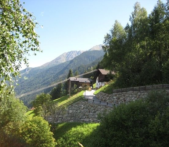 Ein landschaftlicher Blick auf einen Berg mit einem Haus am Hang, umgeben von üppigem Grün und Bäumen. Ein Weg führt zum Haus.