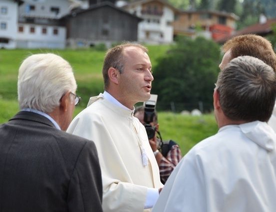 Ein Priester in einer weißen Robe unterhält sich mit zwei Männern, einer mit Brille, in einem Grasfeld mit Häusern im Hintergrund.