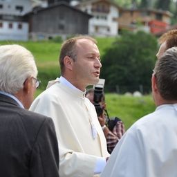 Ein Priester in einer weißen Robe unterhält sich mit zwei Männern, einer mit Brille, in einem Grasfeld mit Häusern im Hintergrund.