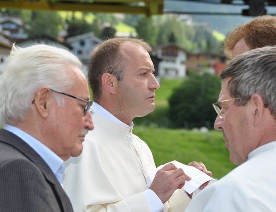 Ein Priester in einer weißen Robe liest aus einem Buch, während drei Männer in Anzügen aufmerksam zuhören, auf einem grünen Feld mit Häusern im Hintergrund.