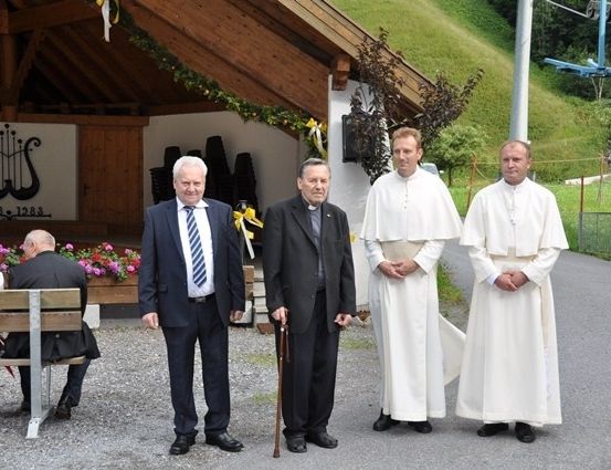 Vier Männer, zwei in Anzügen und zwei in weißen Gewändern, stehen vor einem hölzernen Gebäude mit einem Schild und Blumen. Eine Bank mit Menschen sitzt in der Nähe.