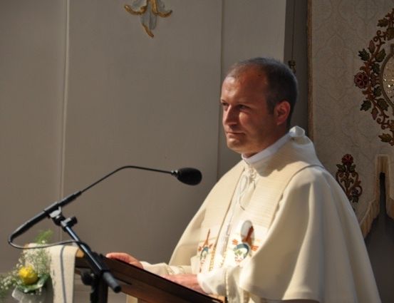Ein Priester steht in einer Kirche an einem Pult und liest aus einem Buch, mit einem Mikrofon und Blumen in der Nähe.