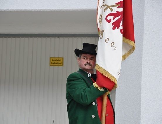 Ein Mann in einem grünen Mantel und schwarzem Hut hält eine Fahne mit rotem Hintergrund und weißem Design. Er steht vor einer weißen Wand mit einem gelben Schild.