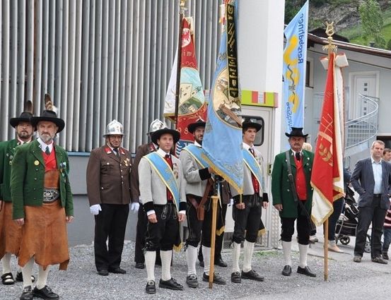 Eine Gruppe von Männern in traditioneller Kleidung, möglicherweise für eine feierliche Veranstaltung, steht in einer Reihe mit Fahnen und Bannern. Sie stehen vor einem Gebäude mit einer Wendeltreppe.