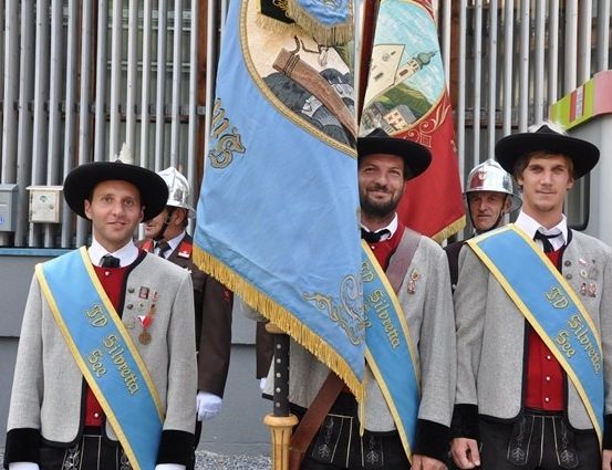Vier Männer in traditionellen Uniformen, einschließlich Hüten und Schärpen, stehen vor einem Gebäude. Sie halten eine Fahne mit einem goldenen Design. Dahinter steht ein weiterer Mann in Helm und Uniform.