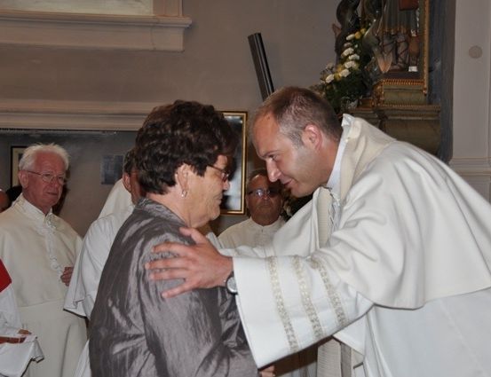 Ein Priester in einer weißen Robe tröstet eine ältere Frau in einem grauen Kleid während einer religiösen Zeremonie.