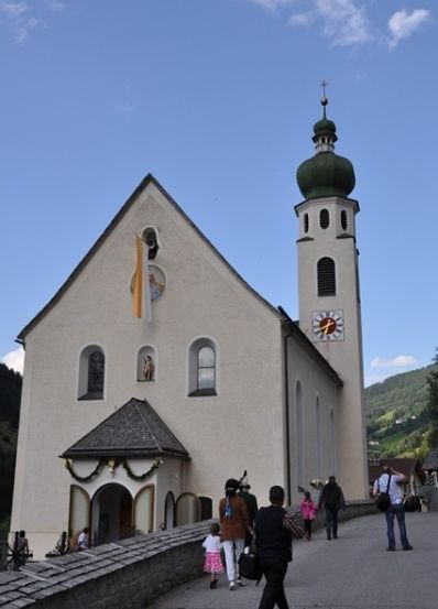 Eine traditionelle Kirche mit einer grünen Kuppel und einem Glockenturm, mit Menschen, die an einem sonnigen Tag davor spazieren.