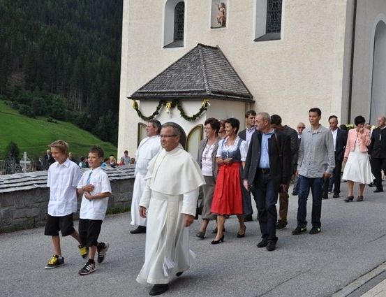 Ein Priester führt eine Prozession von Menschen vor einer Kirche an, mit einem bergigen Hintergrund.