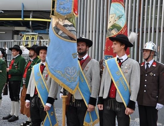 Eine Gruppe von Männern in traditionellen Uniformen, einige halten Fahnen, stehen in einer Reihe vor einem Gebäude.