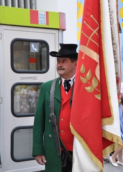 Ein Mann in einem grünen und roten Kostüm mit einem schwarzen Hut steht neben einer roten Fahne. Er hält ein Gewehr und steht vor einer Telefonzelle.