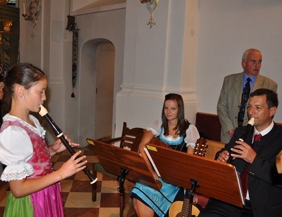 Ein junges Mädchen in traditioneller Kleidung spielt eine Blockflöte, während ein Mann in Anzug neben einer Frau in traditioneller Kleidung mit einer Gitarre eine Blockflöte spielt.