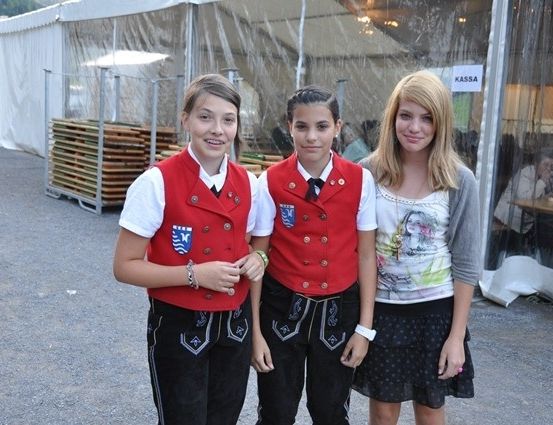 Drei Mädchen stehen zusammen vor einem Zelt, zwei in traditioneller Kleidung und eine in lässiger Kleidung. Hinter ihnen ein verschwommener Blick auf einen Tisch mit sitzenden Personen.