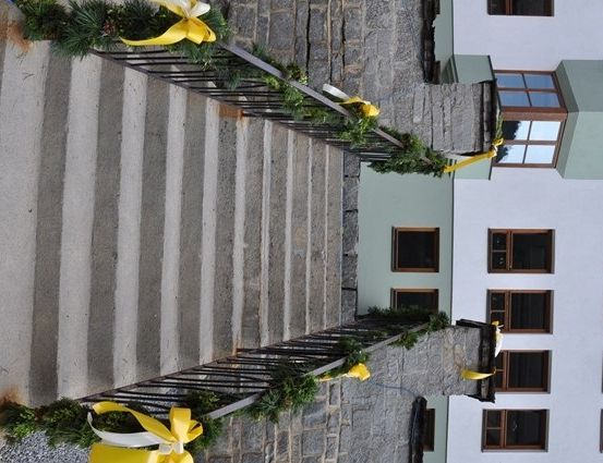 Auf einer Steintreppe schmücken gelbe Bänder die Geländer und führen zu einem Gebäude mit mehreren Fenstern und einem grün umrahmten Eingang.