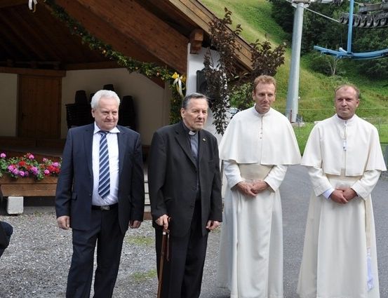 Drei Priester in weißen Gewändern und ein Mann in Anzug stehen vor einem Gebäude mit einem Schild, das 1383 steht.