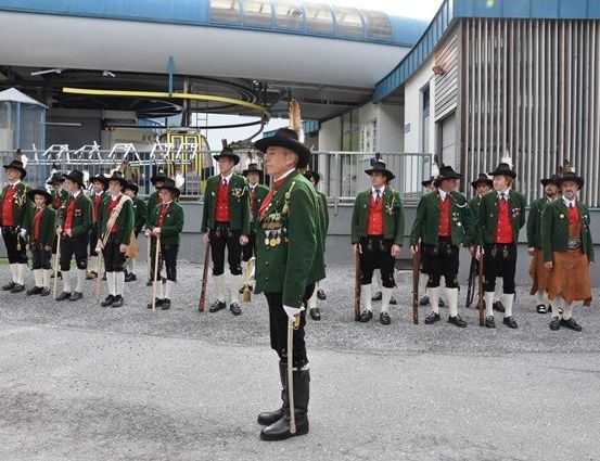 Eine Gruppe von Männern in traditionellen grünen und roten Uniformen steht in einer Reihe und hält Gewehre vor einem Gebäude.