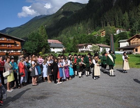 Eine Gruppe von Menschen in traditioneller Kleidung steht in einer Reihe, wahrscheinlich für einen Umzug, vor einem Berg mit Häusern und Bäumen.