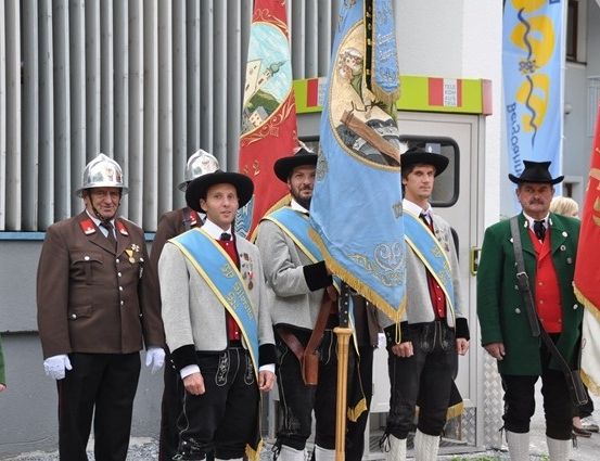 Eine Gruppe von Männern in traditionellen Uniformen steht in einer Reihe und hält Flaggen. Sie tragen Hüte, Medaillen und Lederschuhe. Der Mann in der Mitte hält eine Flagge mit einem goldenen Stab. Hinter ihnen steht ein Gebäude mit einer Glastür.