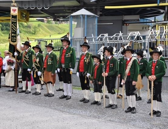 Eine Gruppe von Männern in traditionellen grünen Uniformen und Hüten steht in einer Reihe an einem Bahnhof mit einer Fahne und einer Straßenbahn im Hintergrund.