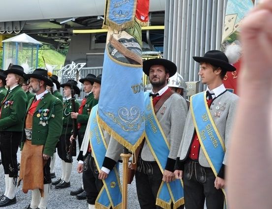 Eine Gruppe von Männern in traditionellen Uniformen, die Gewehre und Fahnen halten, steht in einer Reihe unter einer Metallstruktur. Sie nehmen wahrscheinlich an einer Parade oder einem zeremoniellen Ereignis teil.