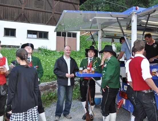 Eine Gruppe von Menschen in traditioneller bayerischer Kleidung versammelt sich um einen blauen Imbisswagen, einige halten Getränke und andere unterhalten sich. Der Hintergrund zeigt ein rustikales Gebäude.