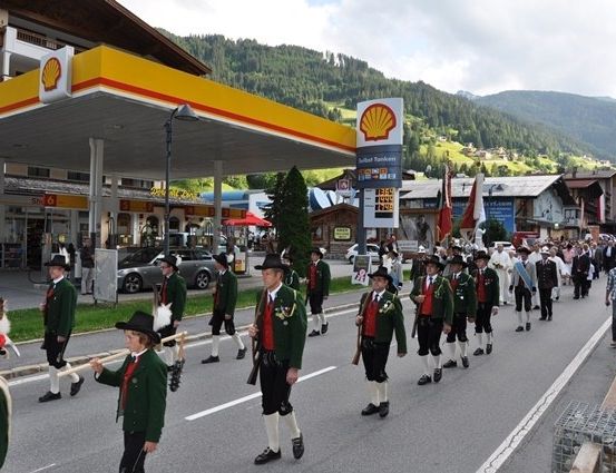 Eine Gruppe von Menschen in traditionellen grünen und weißen Uniformen marschiert eine Straße hinunter, mit einer Shell-Tankstelle im Hintergrund.