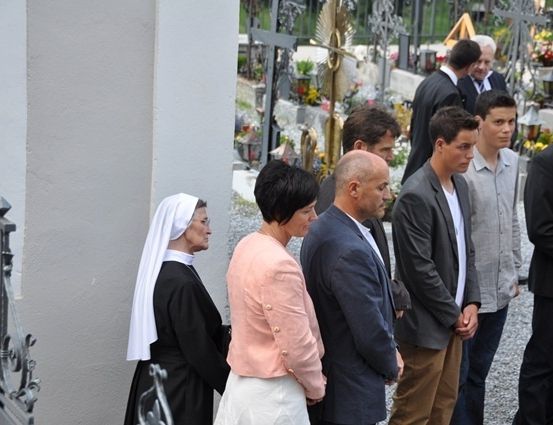 Eine Gruppe von Menschen, darunter eine Nonne, steht auf einem Friedhof mit einer Kulisse von Gräbern und einer Kirche.